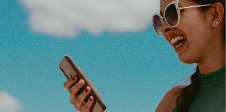Woman smiling at her phone outdoors under a blue sky.
