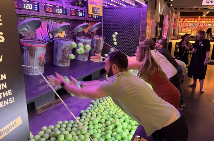 A group of adults playing an arcade-style ball-throwing game, tossing green balls toward targets shaped like trash cans. The setting is lively with bright lights and other players in the background.