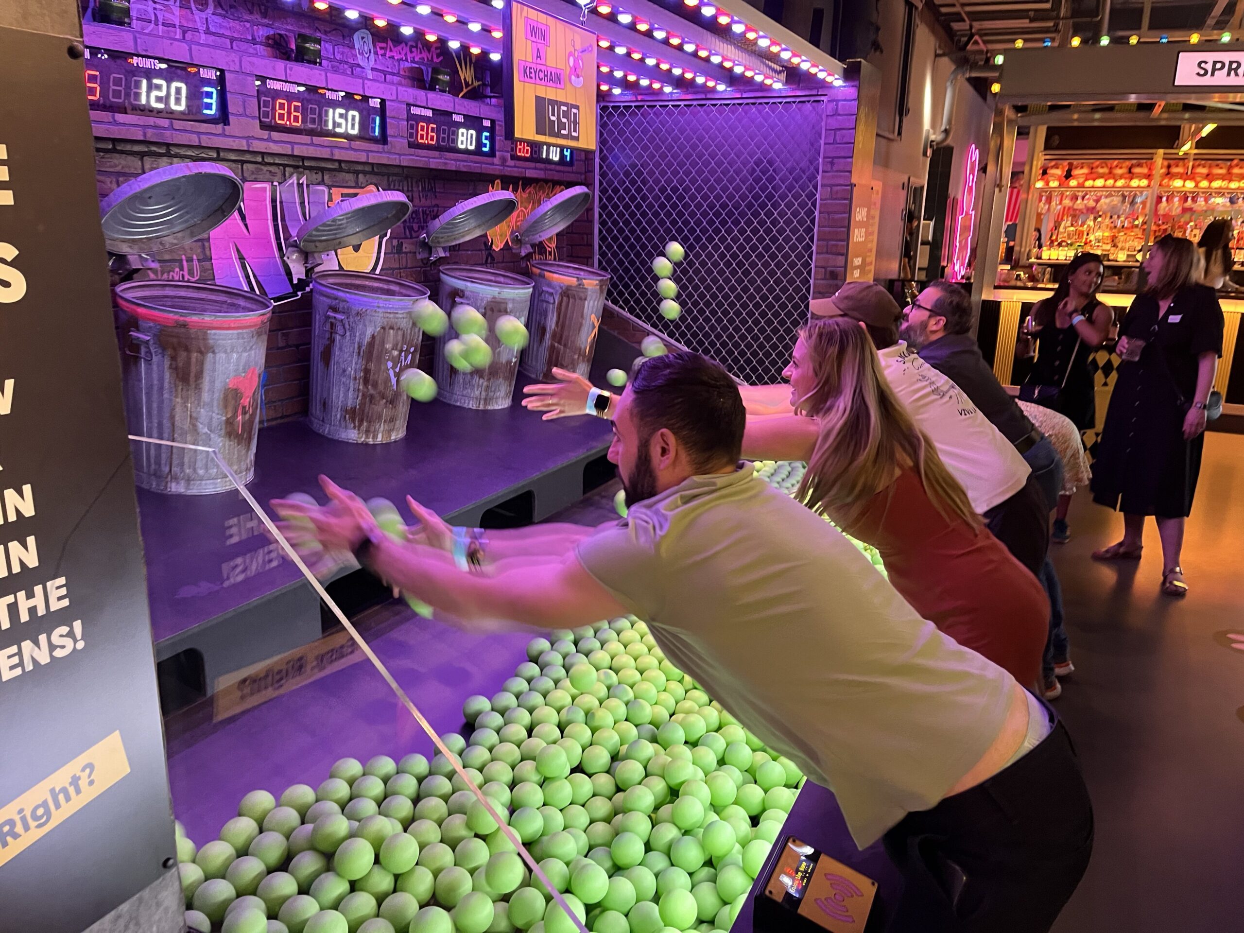 A group of adults playing an arcade-style ball-throwing game, tossing green balls toward targets shaped like trash cans. The setting is lively with bright lights and other players in the background.
