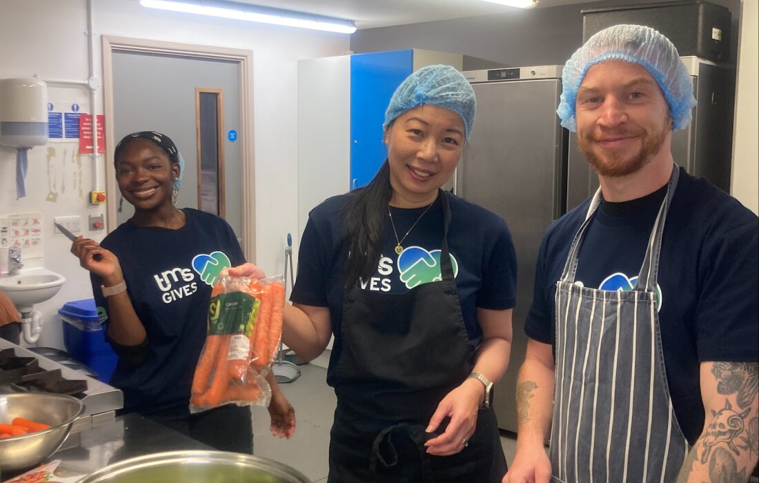 3 people cooking smiling for the camera in hair nets in a kitchen and one woman holding carrots