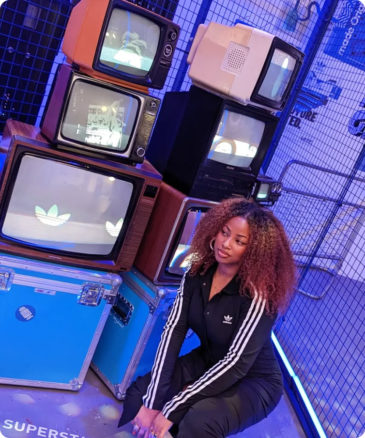A woman in a black Adidas tracksuit crouches beside a stack of vintage TVs showing Adidas logos, lit with blue and purple lighting.