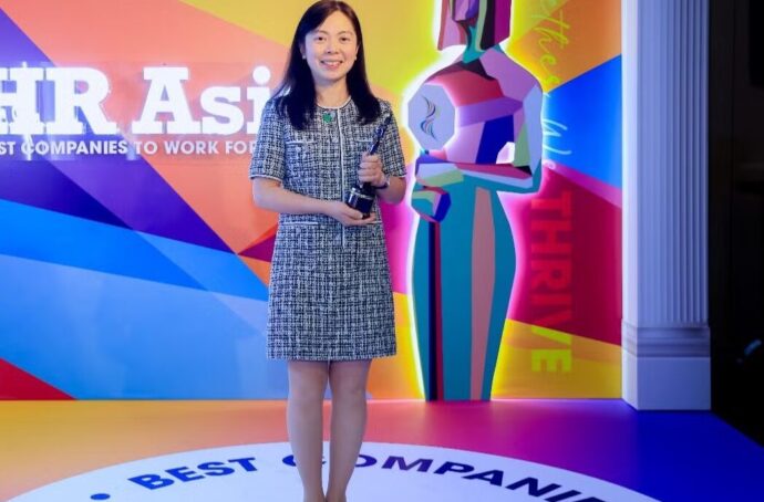 A woman standing on a brightly lit stage holding an award trophy. The backdrop features colorful graphics and the text “HR Asia, Best Companies to Work For.”