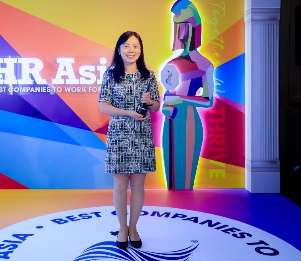 A woman standing on a brightly lit stage holding an award trophy. The backdrop features colorful graphics and the text “HR Asia, Best Companies to Work For.”