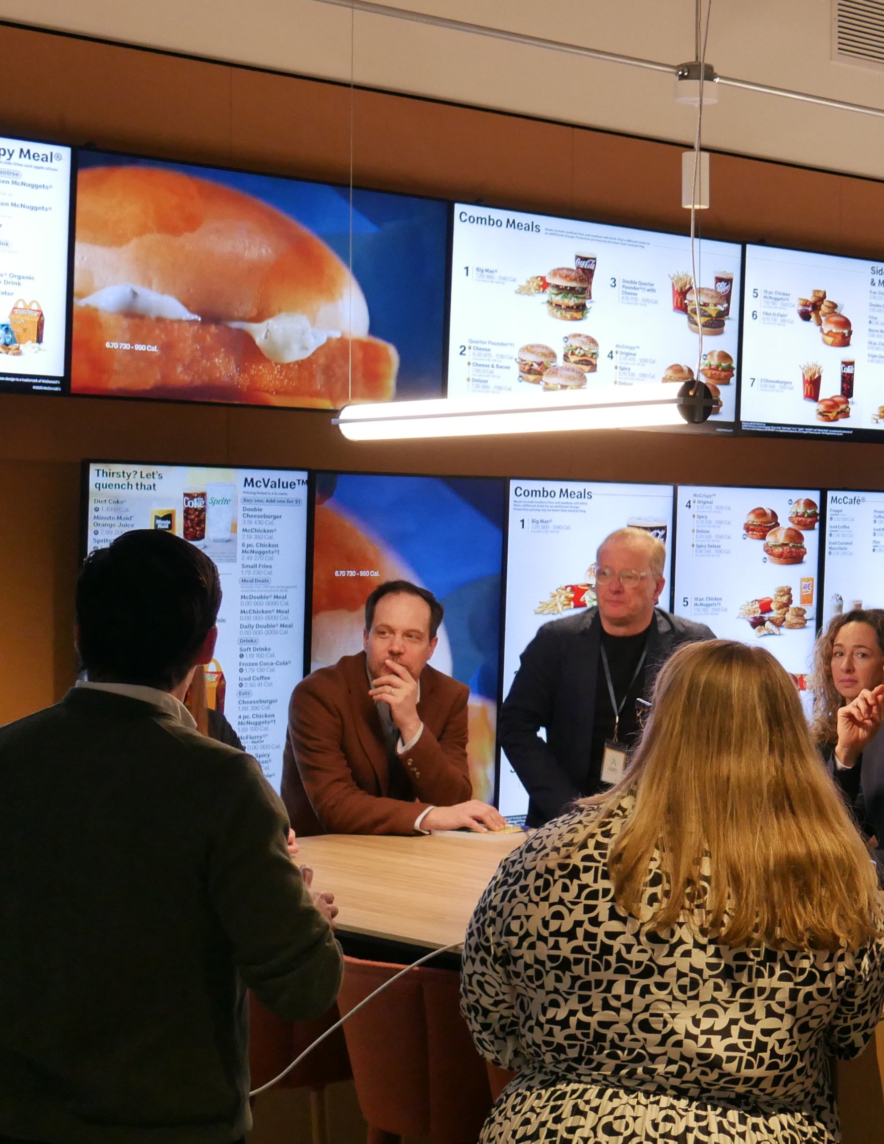 A group of people gathered around a table inside a McDonald’s restaurant. Several digital menu boards are displayed behind them.