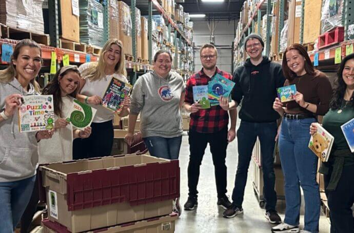 A group of tms volunteers at the Bernie's Book Bank warehouse with books smiling at the camera