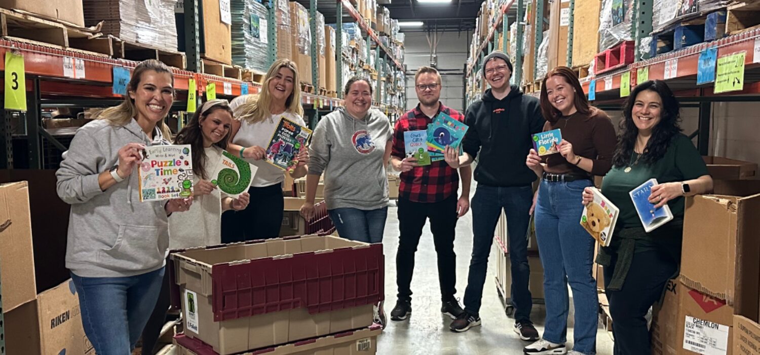 A group of tms volunteers at the Bernie's Book Bank warehouse with books smiling at the camera