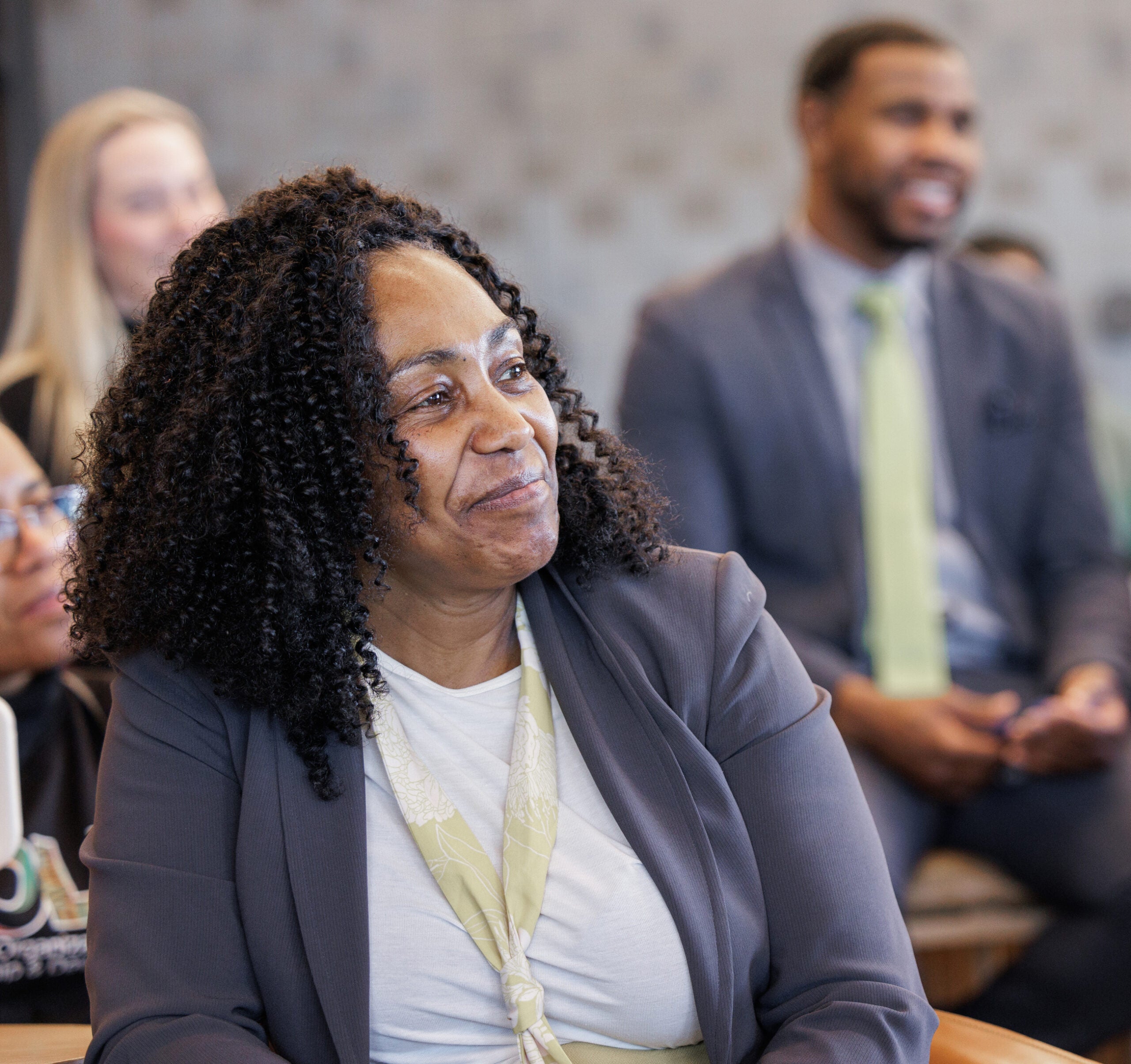 A woman sitting among a group of people, smiling and listening during what appears to be a meeting or presentation.