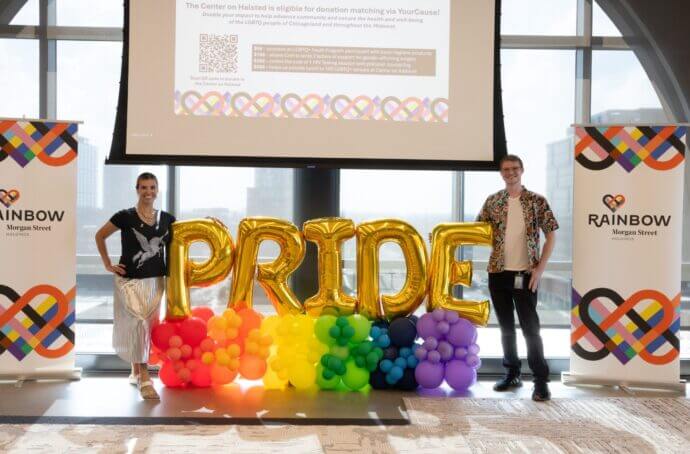 Two people standing beside large gold balloon letters spelling “PRIDE,” arranged above rainbow-colored balloon clusters, with Rainbow Morgan Street branding and a donation-matching presentation displayed in the background.