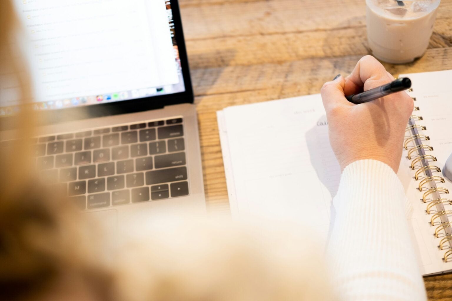 Over-the-shoulder shot of a lady's hand holding a pen, writing in a golden spiral-bound notebook beside an open Apple laptop. There's a half-drunk iced-coffee on the wooden table