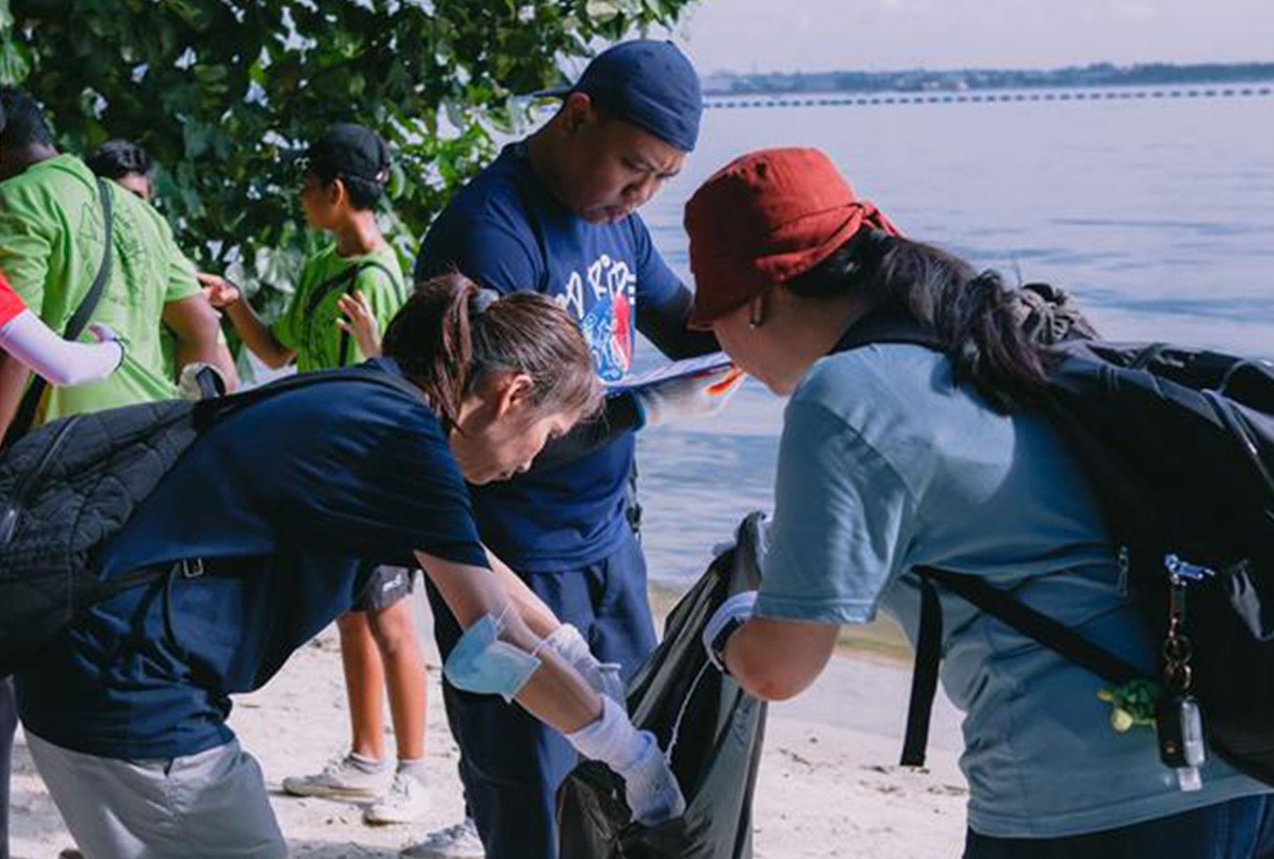 Des personnes travaillant ensemble lors d’un nettoyage de plage, portant des gants et déposant les déchets ramassés dans un grand sac près du rivage.