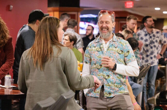 Des personnes discutant lors d’un événement dans un bowling. Un homme en chemise à motifs rit en tenant un verre et en parlant avec un autre participant. 