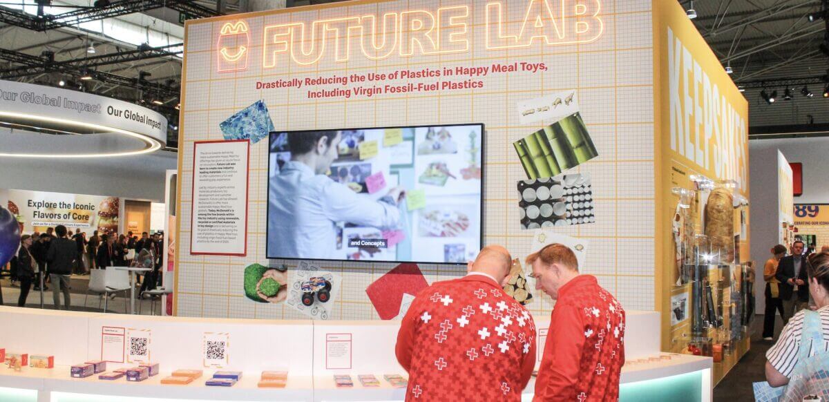A McDonald’s “Future Lab” exhibit at a trade show, featuring a large screen, colorful material samples on the wall, and displays about reducing plastics in Happy Meal toys. Two attendees in matching red jackets are viewing items at the counter, with other visitors walking through the exhibit area.