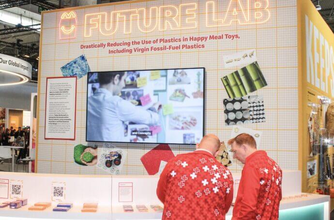 Un stand « Future Lab » de McDonald’s lors d’un salon, avec un grand écran, des échantillons de matériaux colorés au mur et des présentations sur la réduction du plastique dans les jouets Happy Meal. Deux visiteurs en vestes rouges identiques observent les éléments exposés, tandis que d’autres parcourent l’espace. 