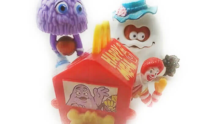 Des figurines de personnages McDonald’s entourent un jouet en forme de boîte Happy Meal rouge. On voit notamment une figurine violette aux grands yeux, une figurine blanche avec un chapeau bleu, et une petite figurine de clown devant la boîte.