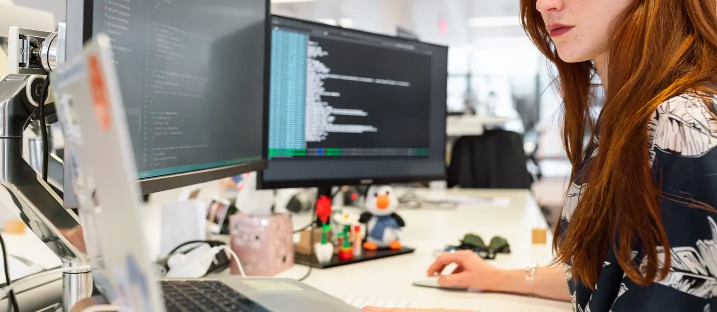 Red-headed woman works on code across three monitors