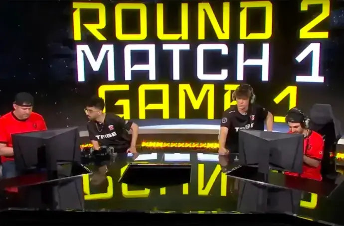 4 gamers behind 2 computer screens and yellow and black background that says "Round 2 Match 1 Game 1"