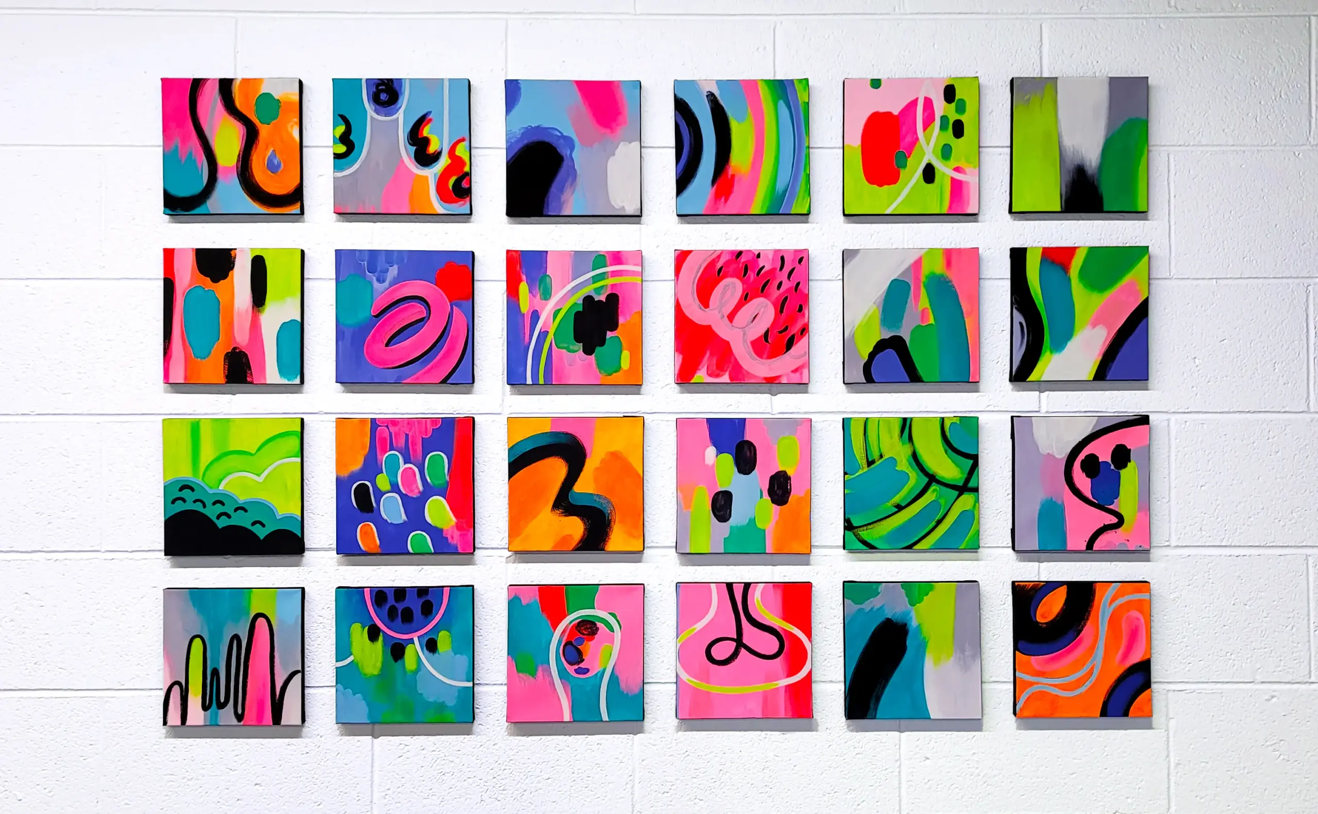 A grid of 24 square canvases with abstract flurescent art, on a white brick wall.