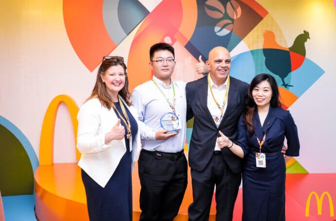 Quatre personnes posant devant un décor coloré sur le thème de McDonald’s. L’une d’elles tient un prix, et le groupe sourit en faisant un signe de pouce levé.