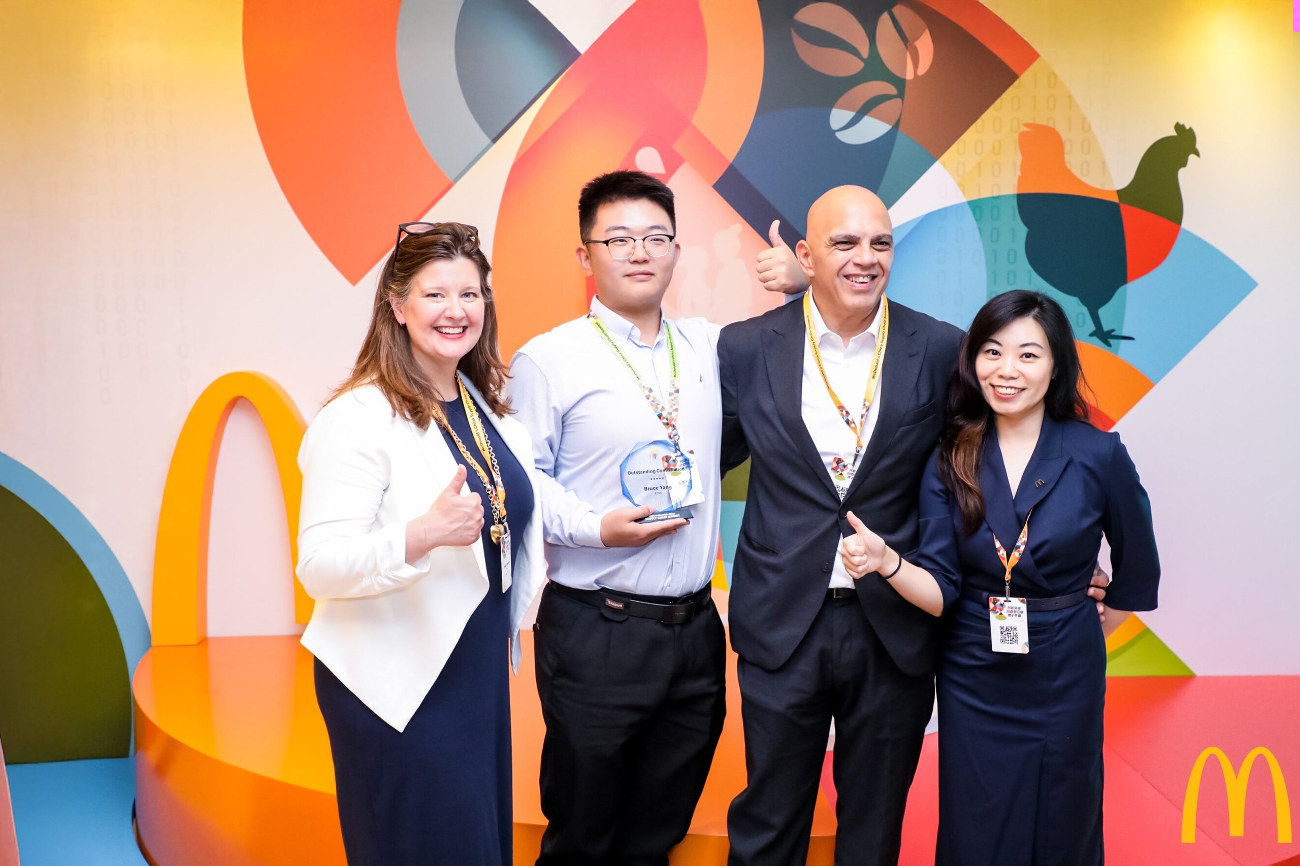 Quatre personnes posant devant un décor coloré sur le thème de McDonald’s. L’une d’elles tient un prix, et le groupe sourit en faisant un signe de pouce levé.