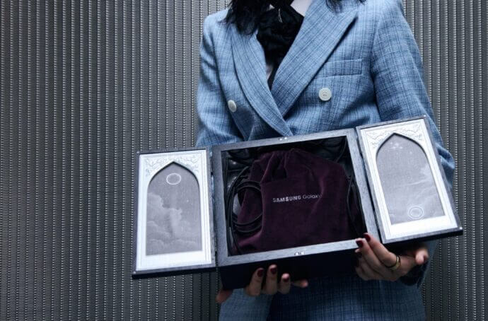 A person in a blue suit holding an open presentation box that displays a velvet Samsung Galaxy pouch, styled with ornate gothic-frame panels on each side.